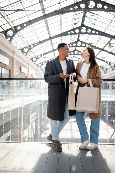 Piena lunghezza della coppia multietnica alla moda con borse della spesa parlando vicino al centro commerciale offuscato — Foto stock