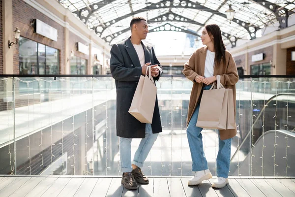 Pleine longueur de couple interracial en manteaux et jeans tenant des sacs à provisions et se regardant près du centre commercial — Photo de stock