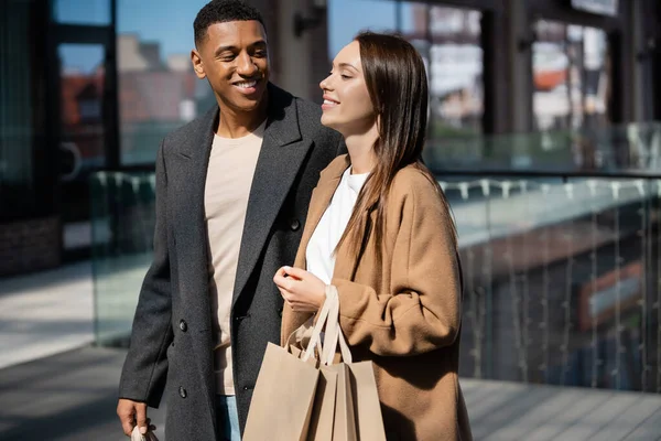 Lächelnder afrikanisch-amerikanischer Mann schaut fröhliche junge Freundin mit Einkaufstüten auf der Straße an — Stockfoto