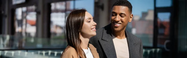 Hombre afroamericano feliz mirando a novia joven y feliz en la calle urbana, bandera - foto de stock