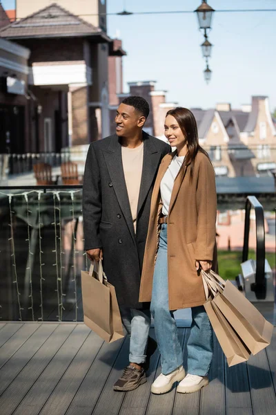In voller Länge multiethnisches Paar in modischen Kleidern mit Einkaufstaschen vor verschwommenem Hintergrund der Stadt — Stockfoto