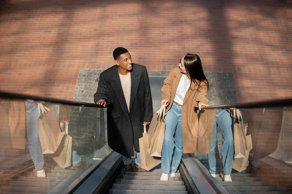 Visão de alto ângulo de casal interracial na moda com sacos de compras sorrindo um para o outro na escada rolante — Fotografia de Stock
