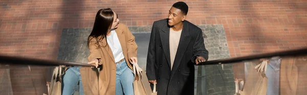 Visão de alto ângulo de casal multiétnico sorridente com sacos de compras olhando uns para os outros na escada rolante, banner — Fotografia de Stock