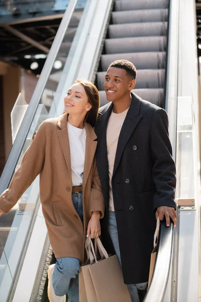 Feliz e elegante casal multiétnico com sacos de compras olhando para longe na escada rolante — Fotografia de Stock