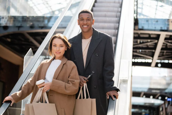Couple interracial en manteaux à la mode tenant des sacs à provisions et regardant la caméra sur l'escalator — Photo de stock