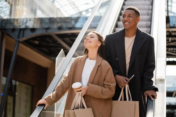 Alegre e elegante casal multiétnico com compras olhando para longe na escada rolante — Fotografia de Stock