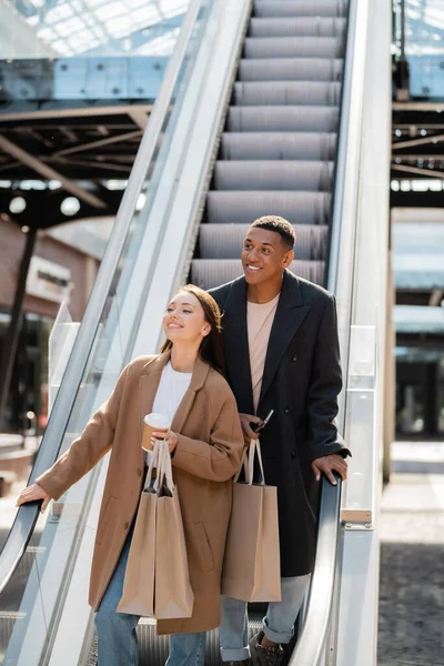 Pareja interracial con estilo con bolsas de compras sonriendo y mirando hacia otro lado en escaleras mecánicas - foto de stock