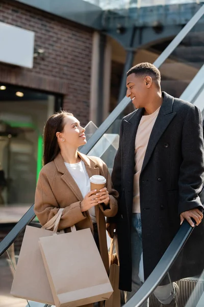 Giovane donna con caffè per andare e shopping bags sorridente alla moda afro americano uomo sulla scala mobile — Foto stock