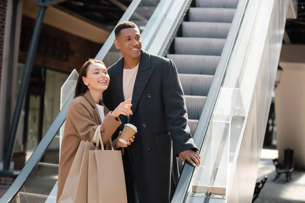 Cheerful woman with paper cup and shopping bags pointing with finger near trendy african american man on escalator — Stock Photo