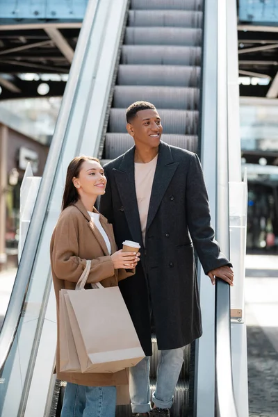 Africano americano hombre en elegante abrigo mirando lejos cerca joven novia con café para ir y bolsas de compras en escaleras mecánicas - foto de stock