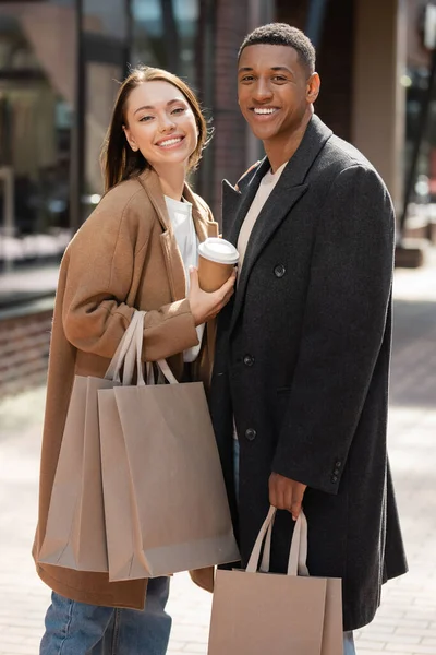 Jovem mulher satisfeita com bebida takeaway e sacos de compras olhando para a câmera perto de homem americano africano na moda — Fotografia de Stock