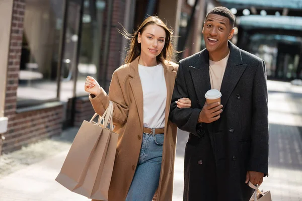 Divertido y alegre afroamericano hombre mirando a la cámara cerca de novia joven con bolsas de compras - foto de stock