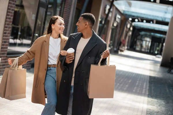 Trendy multiethnic couple in coats holding shopping bags and smiling at each other on urban street — Stock Photo
