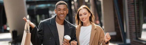 Alegre hombre afroamericano con bolsa de compras y taza de papel cerca de feliz novia joven, pancarta - foto de stock