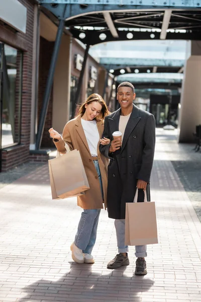 Élégant homme afro-américain avec des sacs à provisions et tasse en papier regardant la caméra près de petite amie gaie — Photo de stock