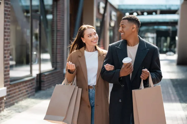 Heureux couple multiethnique en vêtements à la mode en se regardant tout en portant des sacs à provisions dans la rue — Photo de stock