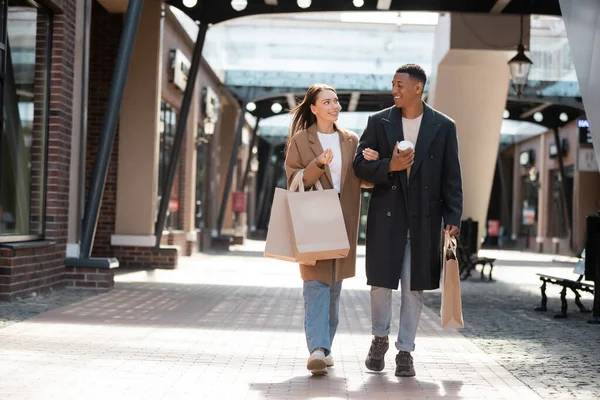 Mulher sorridente com sacos de compras conversando com o namorado afro-americano enquanto caminhava na rua da cidade — Fotografia de Stock