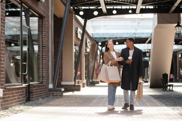 Happy interracial couple in trendy coats walking with purchases along shops on urban street — Stock Photo