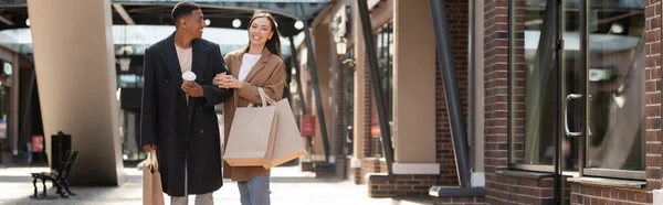 Pareja multiétnica de moda con bolsas de compras y taza de papel sonriendo cerca de tiendas en la calle, pancarta - foto de stock