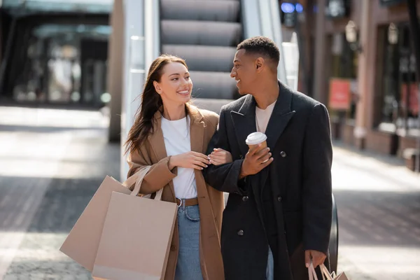 Cheerful and stylish multiethnic couple with shopping bags and takeaway drink looking at each other on urban street — Stock Photo