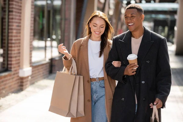 Interracial couple in trendy coats smiling while walking in city with shopping bags and coffee to go — Stock Photo
