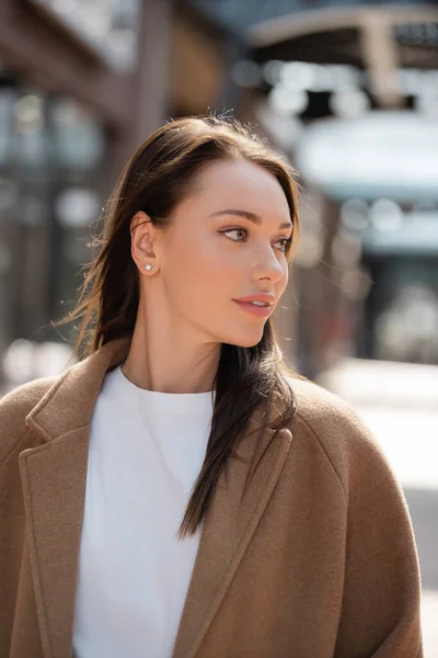 Portrait of pretty brunette woman in beige coat looking away on blurred city street — Stock Photo