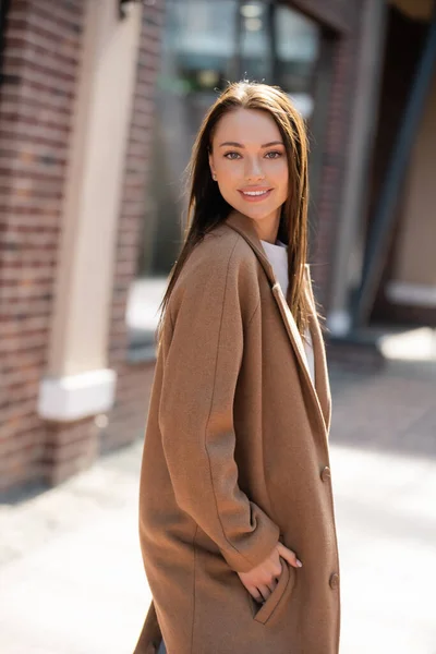 Giovane e bella donna bruna in piedi con mano in tasca di cappotto beige e sorridente alla macchina fotografica — Foto stock