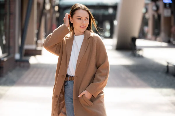 Young woman standing with hand in pocket of beige coat and looking away on blurred street — Stock Photo