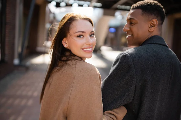 Mulher bonita e feliz olhando para a câmera perto de namorado americano africano na rua turva — Fotografia de Stock