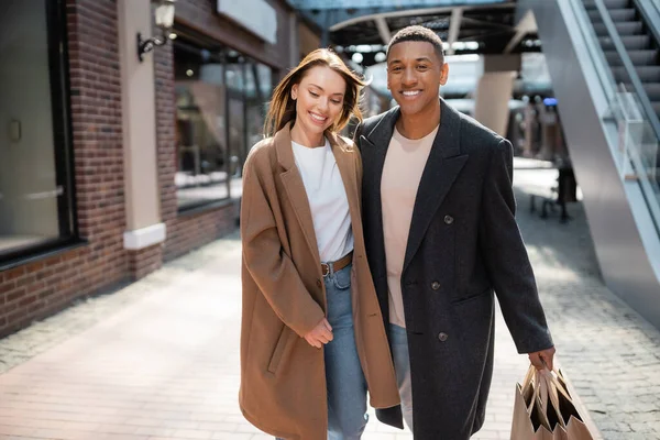 Африканский американец с сумками, смотрящий в камеру рядом с счастливой девушкой в модном пальто — стоковое фото