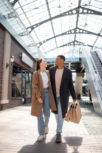 Full length of fashionable interracial couple in coats smiling at each other while walking with shopping bags on street — Stock Photo