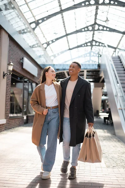 Longitud completa de la mujer elegante sonriendo al novio afroamericano con bolsas de compras cerca del centro comercial - foto de stock