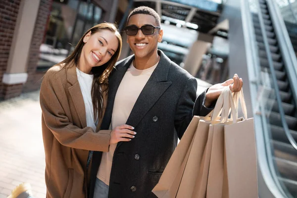 Mulher feliz e elegante abraçando o namorado afro-americano em óculos de sol segurando sacos de compras na rua — Fotografia de Stock