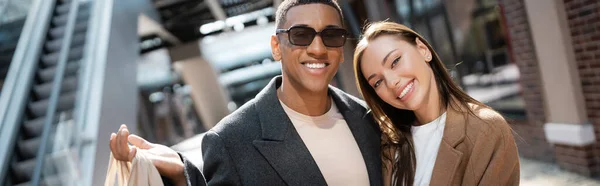 Hombre afroamericano de moda en gafas de sol y bonita mujer joven sonriendo a la cámara al aire libre, pancarta - foto de stock