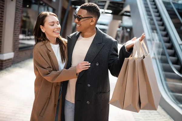 African american man in sunglasses holding shopping bags near happy girlfriend on street — Stock Photo