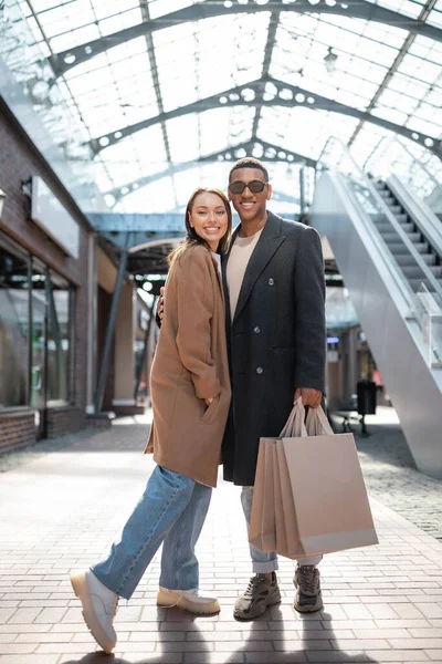 На всю длину улыбающийся африканский американец в солнечных очках, держащий сумки с покупками и обнимающий модную подружку — стоковое фото