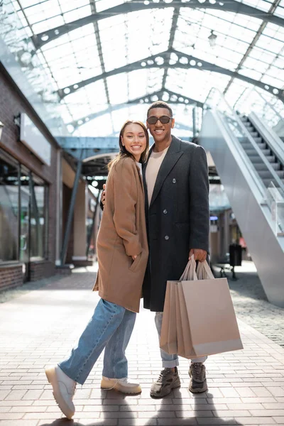 Full length of smiling african american man in sunglasses holding shopping bags and hugging trendy girlfriend — Stock Photo