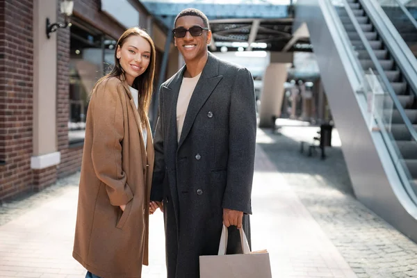 Young woman standing with hand in pocket of coat and holding hands with trendy african american man with shopping bag — Stock Photo