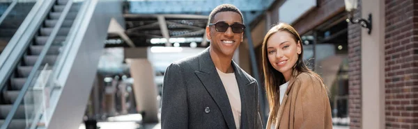 Feliz casal multiétnico em casacos da moda sorrindo para a câmera na rua urbana, banner — Fotografia de Stock