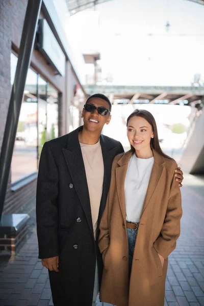 Glücklich afrikanisch-amerikanischer Mann mit Sonnenbrille umarmt junge und stilvolle Freundin auf der Straße der Stadt — Stockfoto