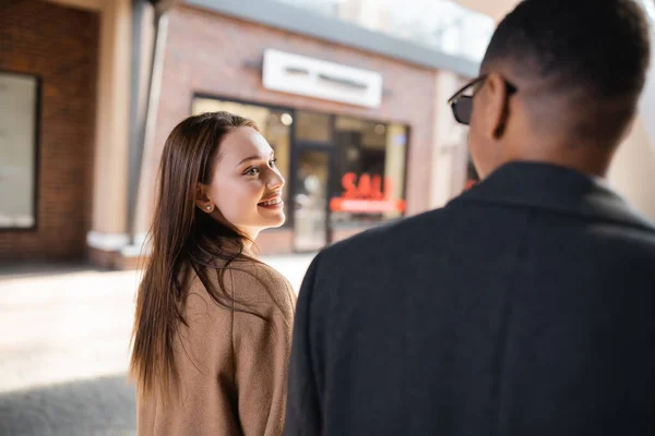 Happy brunette woman looking at african american boyfriend on blurred foreground in city — Stock Photo