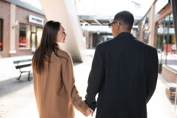 Vista trasera de la pareja multiétnica de moda en abrigos tomados de la mano y mirándose unos a otros en la calle urbana - foto de stock