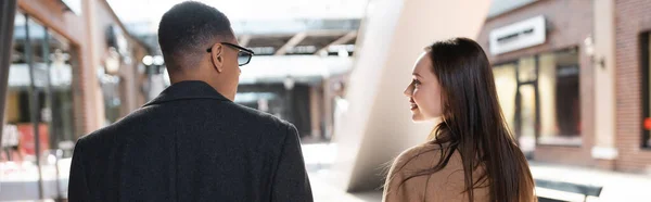 Back view of interracial couple in coats looking at each other on street in city, banner — Stock Photo