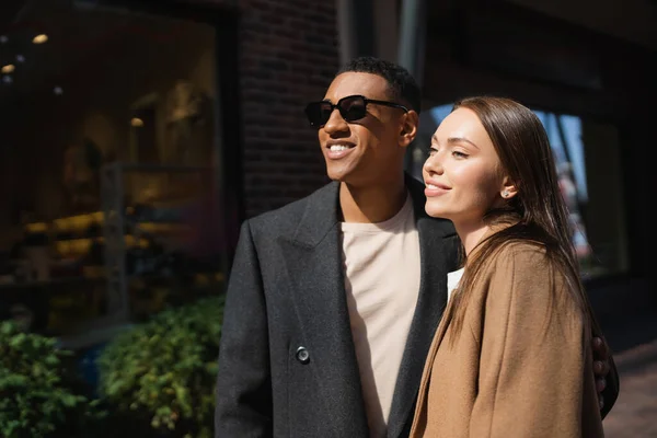 Alegre pareja interracial en abrigos de moda mirando hacia otro lado en la calle urbana - foto de stock