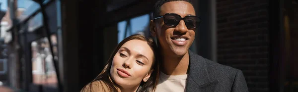 Jovem mulher alegre apoiando-se no homem americano africano feliz em óculos de sol e olhando para a rua, banner — Fotografia de Stock