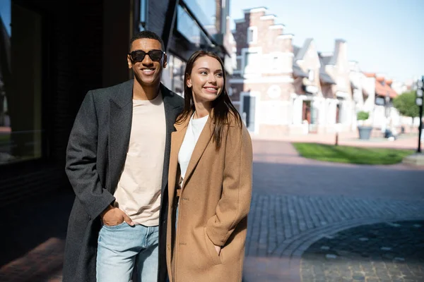 Feliz y de moda pareja multiétnica en abrigos de pie con las manos en los bolsillos en la calle de la ciudad - foto de stock