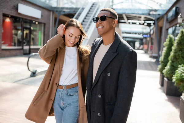 Hombre afroamericano con estilo en gafas de sol sonriendo cerca de novia fijación de pelo en la calle - foto de stock