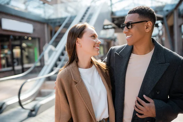 Pareja multiétnica de moda en abrigos sonriendo el uno al otro en la calle urbana borrosa - foto de stock