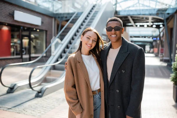 Trendy interracial couple in coats smiling at camera near blurred escalator on street — Stock Photo