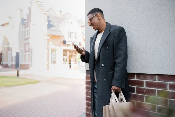Homme afro-américain joyeux en manteau et lunettes de soleil tenant des sacs à provisions et bavardant sur le téléphone portable près du bâtiment — Photo de stock